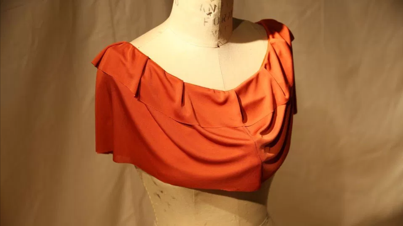 Photo of the front of the wearable piece. The orange ruffled shirt neck is placed on a mannequin. The fabric is deformed on the front where the biting denture toy is placed underneath it.
